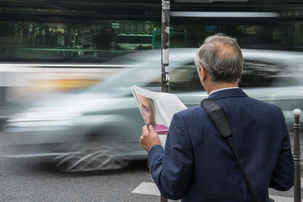 Older man reads a newspaper on a bustling city street with blurred traffic motion.